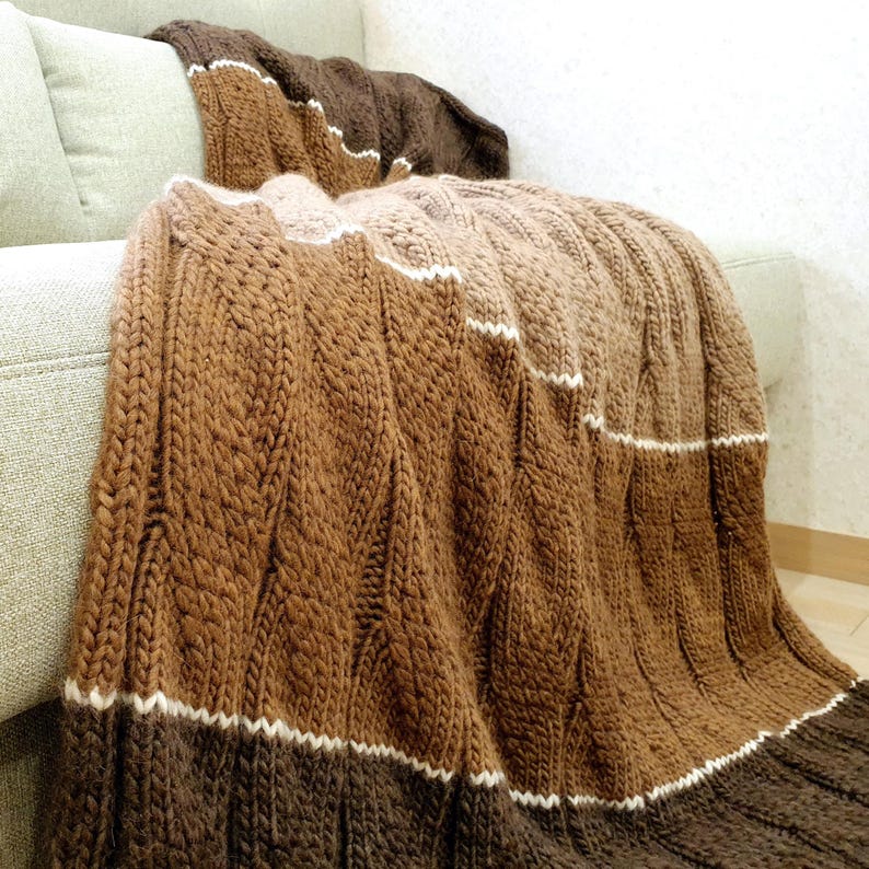 A brown, beige, and white striped knitted throw blanket with a cable knit pattern. The blanket is draped over a couch.