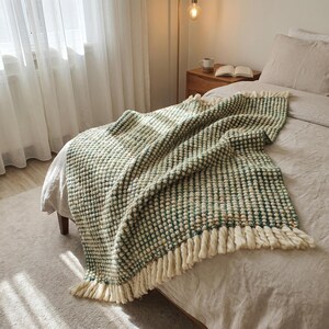 A bed with a linen bedding in Japandi style bedroom. A green and white-colored chunky wool throw blanket with fringe is draped over the bed.
