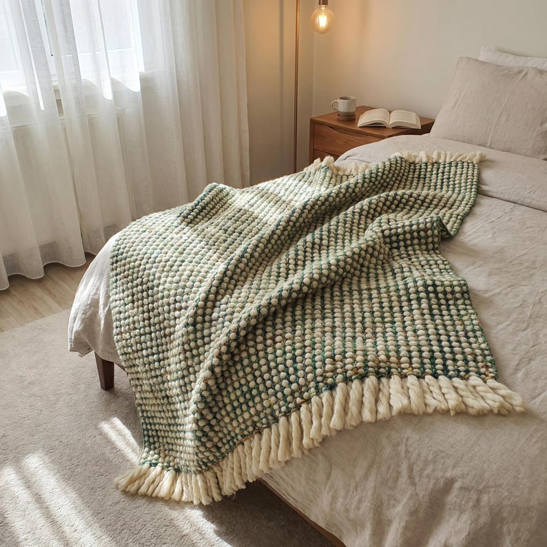 A bed with a linen bedding in Japandi style bedroom. A green and white-colored chunky wool throw blanket with fringe is draped over the bed.