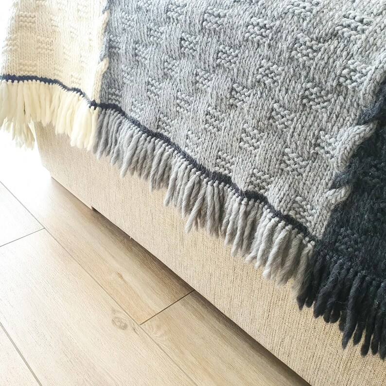 A gray and black knitted throw blanket with fringe on the edges. The blanket is draped over a light beige couch.
