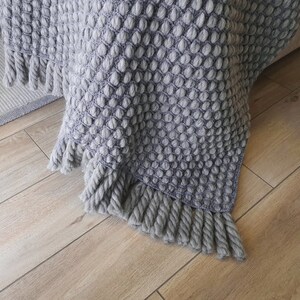 Peut inclure: Une couverture grise tricot&eacute;e avec un motif textur&eacute; en forme de bulles. La couverture a une frange le long du bord inf&eacute;rieur. Le jet&eacute; est drap&eacute; sur une surface, avec un aper&ccedil;u d'un tapis en arri&egrave;re-plan. La couleur est un gris doux et neutre.
