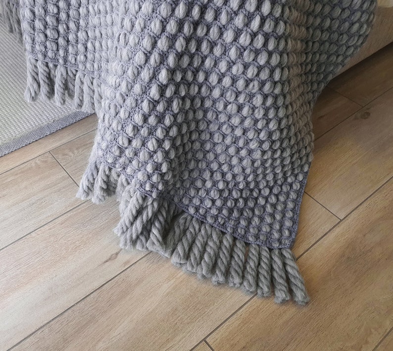 Peut inclure: Une couverture grise tricot&eacute;e avec un motif textur&eacute; en forme de bulles. La couverture a une frange le long du bord inf&eacute;rieur. Le jet&eacute; est drap&eacute; sur une surface, avec un aper&ccedil;u d'un tapis en arri&egrave;re-plan. La couleur est un gris doux et neutre.