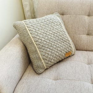 May include: A square, light grey knitted cushion with a woven pattern. The cushion has a cream-coloured border and a small, rectangular tag. The cushion is resting on a beige sofa.