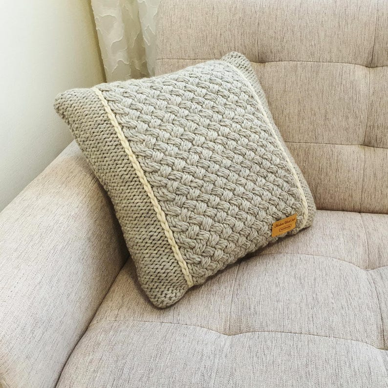 May include: A square, light grey knitted cushion with a woven pattern. The cushion has a cream-coloured border and a small, rectangular tag. The cushion is resting on a beige sofa.