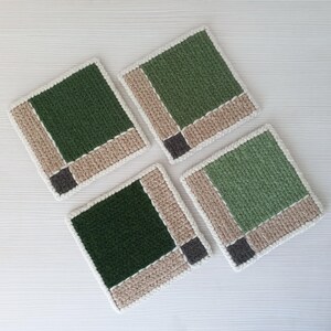 May include: Four square crochet coasters. Each coaster features a dark green center, bordered by tan and white yarn, with a small gray square in the corner. The coasters are arranged on a white surface.