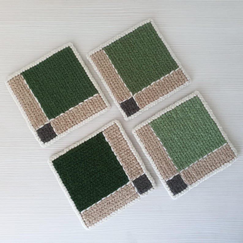 May include: Four square crochet coasters. Each coaster features a dark green center, bordered by tan and white yarn, with a small gray square in the corner. The coasters are arranged on a white surface.