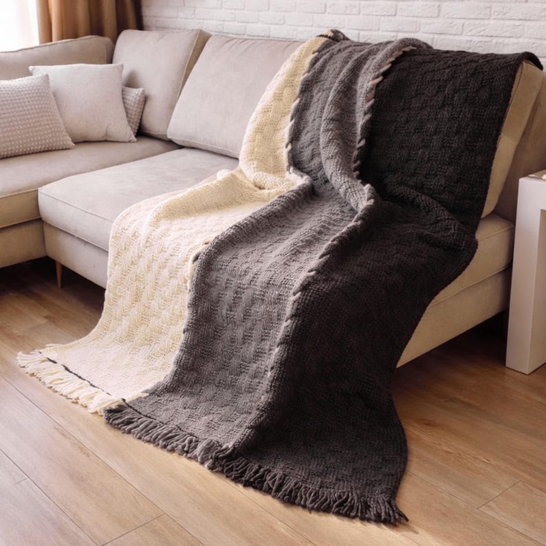 A woven throw blanket with three sections in cream, gray, and black. The blanket has a textured weave and fringe on the edges. The blanket is draped over a couch.