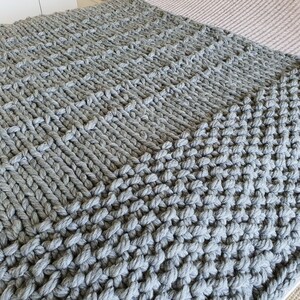 A chunky knit blanket in a soft gray color. The blanket features a textured pattern created by large, thick yarn stitches. The knitted design is visible across the entire surface, creating a cozy and inviting look. The blanket is folded, showing its full size.