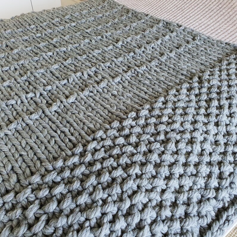 A chunky knit blanket in a soft gray color. The blanket features a textured pattern created by large, thick yarn stitches. The knitted design is visible across the entire surface, creating a cozy and inviting look. The blanket is folded, showing its full size.