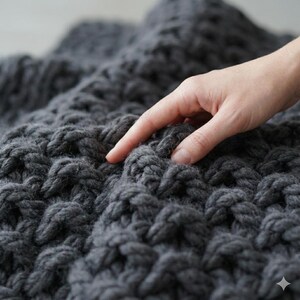 May include: Close-up of a dark gray, chunky knit blanket. The blanket has a textured, raised pattern, and a hand is gently touching the surface. The yarn appears thick and soft, suggesting a cozy, handmade item. The blanket is a solid color.