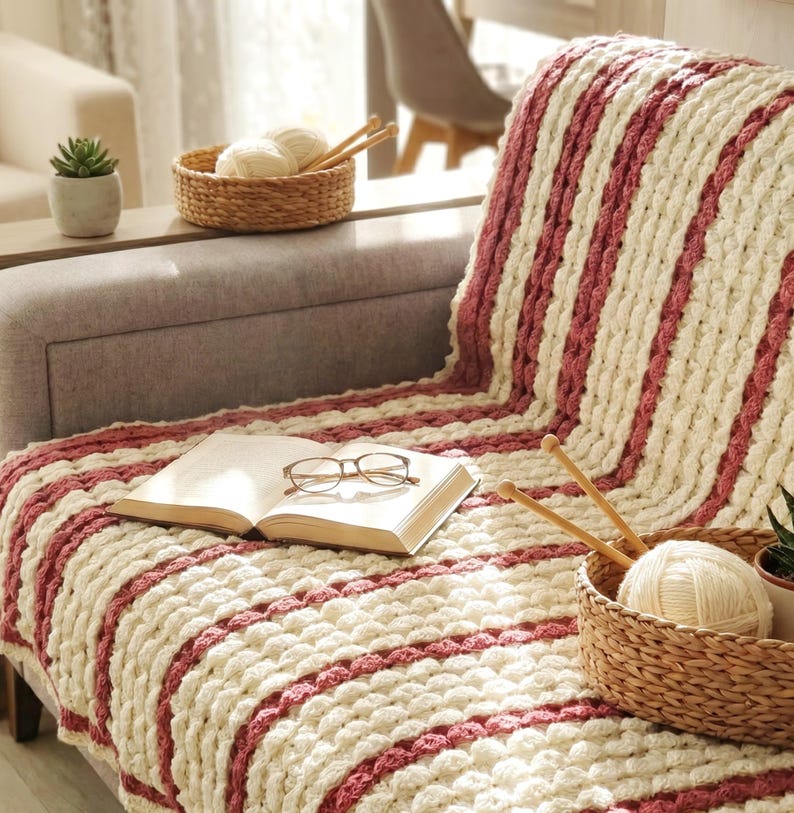 A white and burgundy striped crocheted throw blanket with a textured, bubble design. The blanket is draped over a couch.