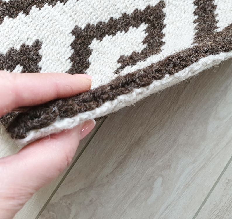 May include: Close-up of a brown and white patterned rug with a fringe edge. The rug is folded in half and being held by a hand.