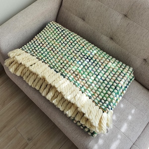 A handmade, textured blanket with a green, white, and blue pattern. The blanket features a cream-colored fringe along the edge. The blanket is folded and resting on a gray sofa.