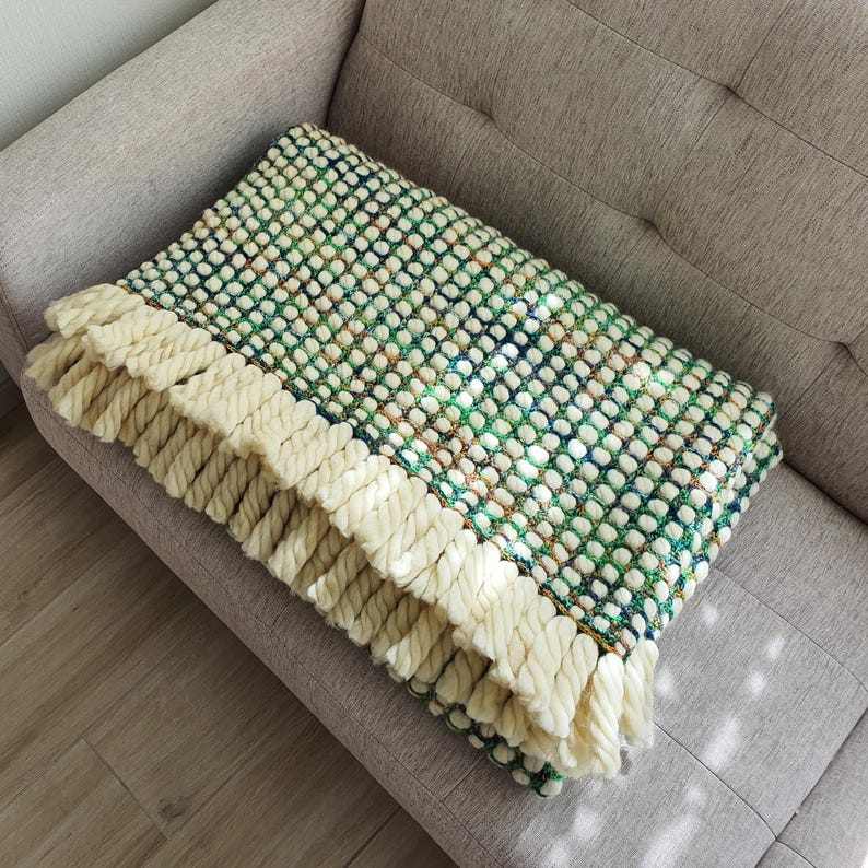 A handmade, textured blanket with a green, white, and blue pattern. The blanket features a cream-colored fringe along the edge. The blanket is folded and resting on a gray sofa.