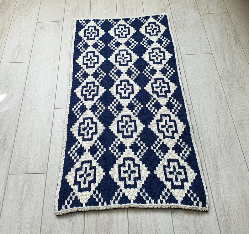May include: A navy blue and white crocheted rug with a geometric pattern. The rug has a diamond shape with a white cross in the center of each diamond.