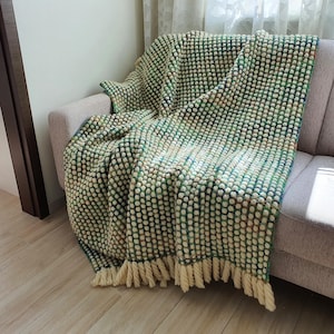 A textured, green and cream-colored throw blanket with a bobble stitch design, draped over a gray sofa. The blanket has a fringe along the bottom edge, adding a decorative touch. The color palette includes shades of green, cream, and a hint of blue.
