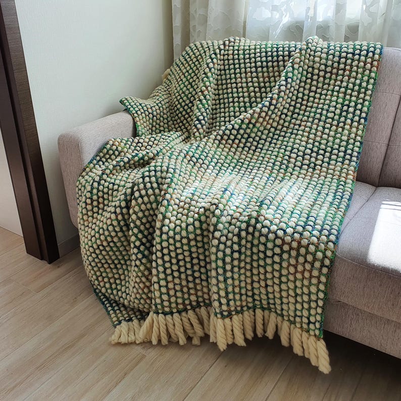 A textured, green and cream-colored throw blanket with a bobble stitch design, draped over a gray sofa. The blanket has a fringe along the bottom edge, adding a decorative touch. The color palette includes shades of green, cream, and a hint of blue.