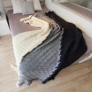 A white, gray, and black knitted throw blanket with fringe on the edges. The blanket is folded and draped over a bed.