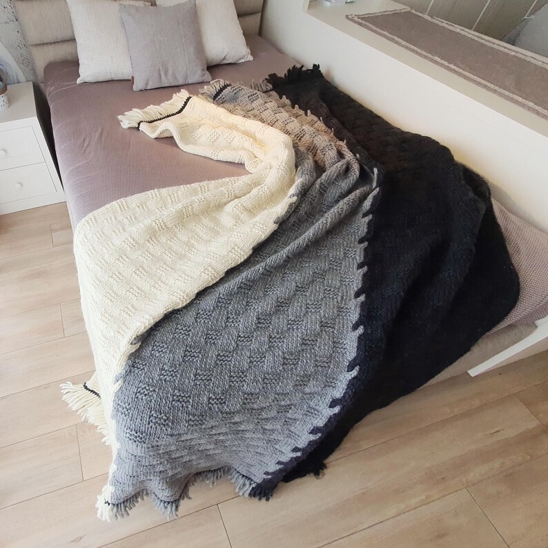 A white, gray, and black knitted throw blanket with fringe on the edges. The blanket is folded and draped over a bed.