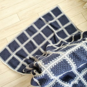 May include: A blue and white crocheted throw blanket with a square pattern. The blanket is folded and draped over a wooden floor.