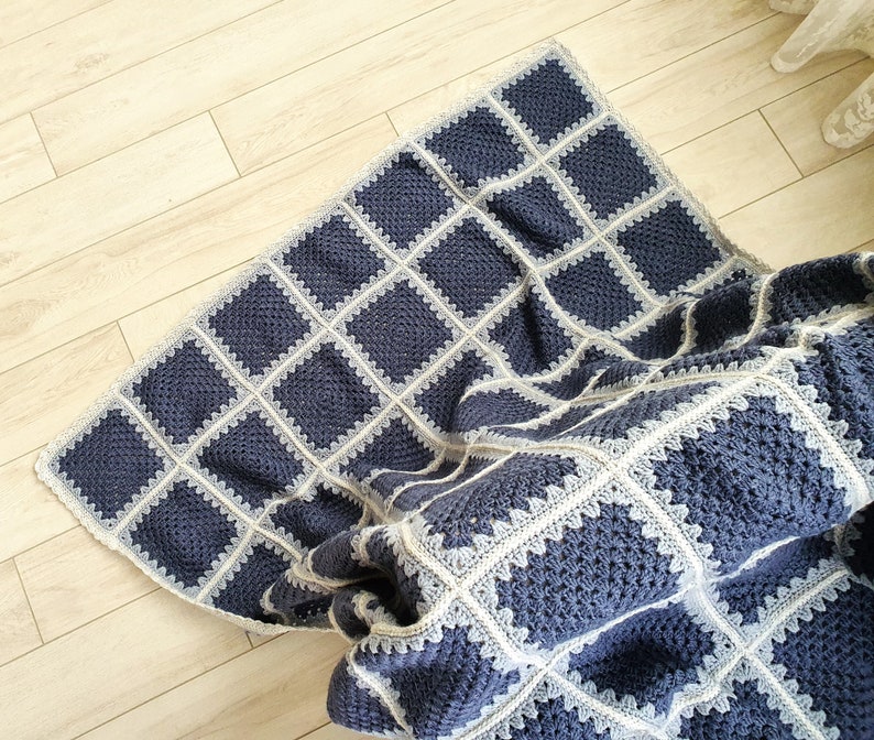 May include: A blue and white crocheted throw blanket with a square pattern. The blanket is folded and draped over a wooden floor.