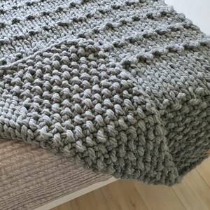 A chunky knit blanket in a soft gray color. The blanket has a textured, raised pattern and is folded over a bed. The thick yarn creates a cozy and inviting look, perfect for home decor and comfort.