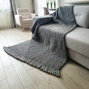 Peut inclure: Une couverture grise en tricot &eacute;pais drap&eacute;e sur un canap&eacute; gris. La couverture pr&eacute;sente un motif textur&eacute; en relief et un bord &agrave; franges. Le canap&eacute; est dans un salon avec une palette de couleurs neutres.