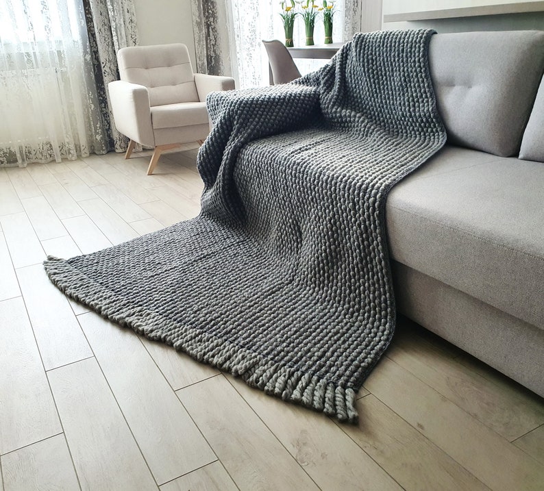 Peut inclure: Une couverture grise en tricot &eacute;pais drap&eacute;e sur un canap&eacute; gris. La couverture pr&eacute;sente un motif textur&eacute; en relief et un bord &agrave; franges. Le canap&eacute; est dans un salon avec une palette de couleurs neutres.