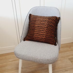 Puede incluir: Un coj&iacute;n cuadrado decorativo marr&oacute;n y naranja con un patr&oacute;n de panal y bordes con flecos. Est&aacute; colocado sobre una silla gris con patas blancas, frente a una pared blanca. Dimensiones aproximadas: 40 cm x 40 cm.