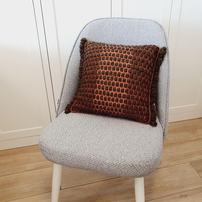Puede incluir: Un coj&iacute;n cuadrado decorativo marr&oacute;n y naranja con un patr&oacute;n de panal y bordes con flecos. Est&aacute; colocado sobre una silla gris con patas blancas, frente a una pared blanca. Dimensiones aproximadas: 40 cm x 40 cm.
