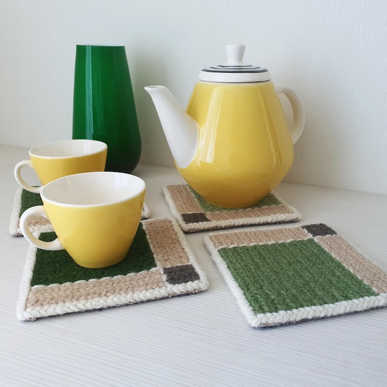 May include: A yellow teapot and two yellow teacups sit on green and beige coasters. A tall green vase is to the left of the teapot. The coasters have a green center with beige and brown borders. The teapot has a white spout and handle.