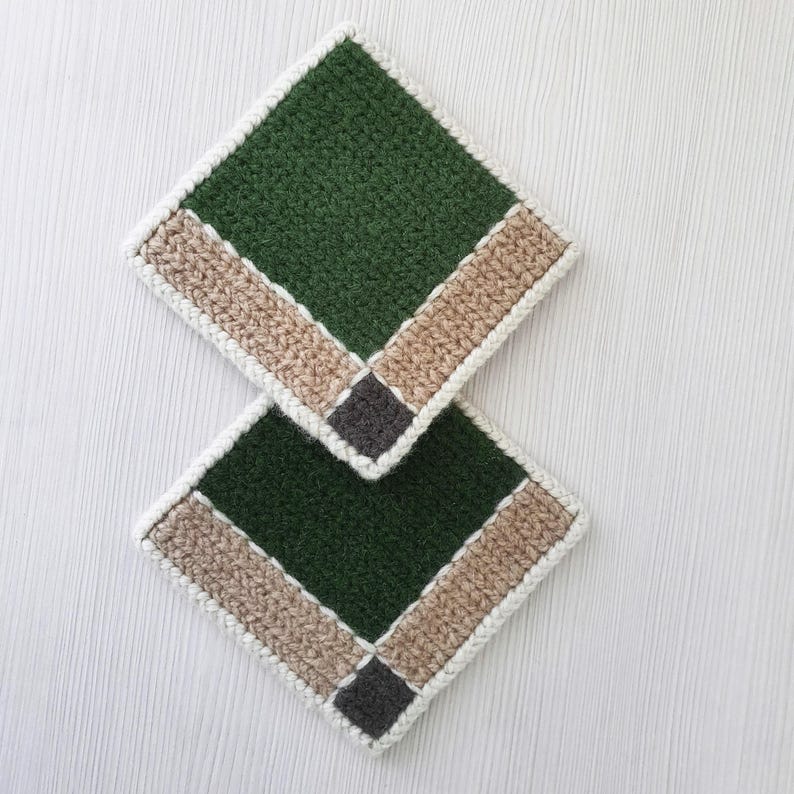 May include: Two handmade, square crochet coasters. Each coaster features a dark green center, bordered by a tan stripe and a white edge. A small gray square accents the corner. The coasters are displayed on a light wood surface.