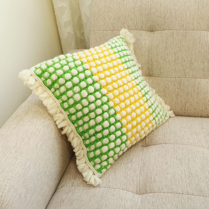 May include: A square decorative pillow with a textured, dotted pattern in green, yellow, and white. The pillow has a fringe trim and is resting on a beige sofa. The pillow's design is divided diagonally with green and yellow sections.