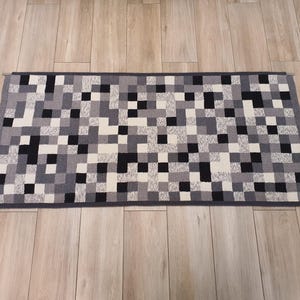 K&ouml;nnte beinhalten: Ein schwarzer, wei&szlig;er und grauer L&auml;ufer mit geometrischem Muster. Der L&auml;ufer ist aus einem weichen, gewebten Material gefertigt und hat einen dezenten Glanz.
