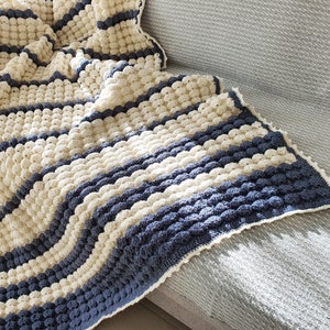 May include: A crocheted throw blanket with a striped pattern in cream and blue. The blanket is made with a textured stitch that creates a puffy, bobbly effect.
