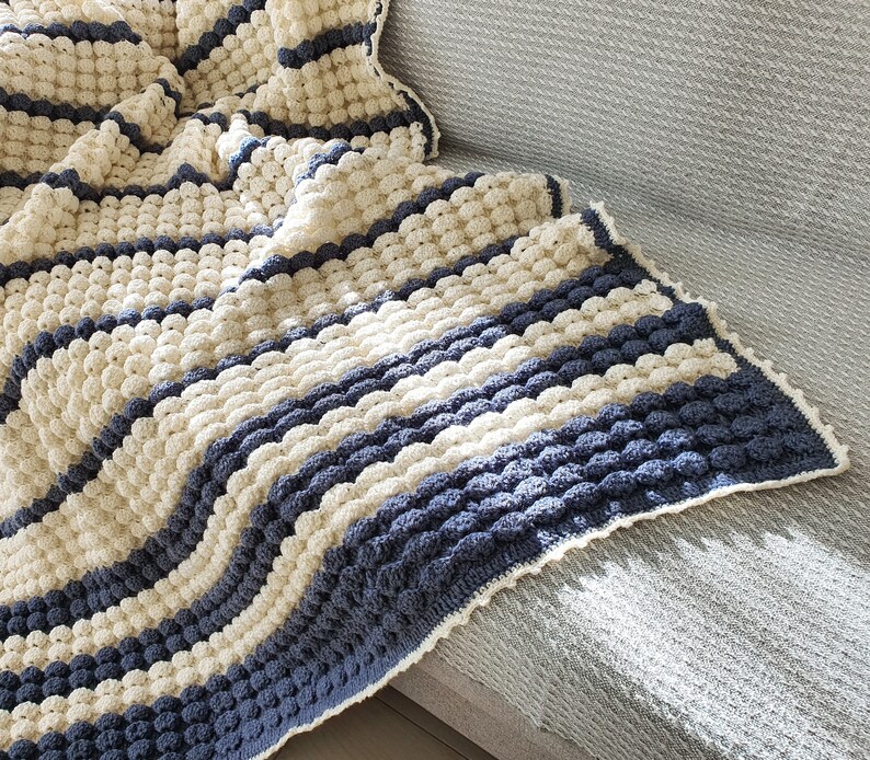 May include: A crocheted throw blanket with a striped pattern in cream and blue. The blanket is made with a textured stitch that creates a puffy, bobbly effect.