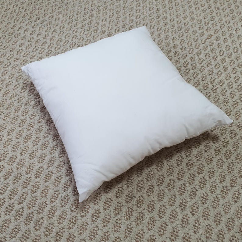K&ouml;nnte beinhalten: Ein quadratisches, wei&szlig;es Kissen auf einem gemusterten beigefarbenen Teppich. Das Kissen ist flauschig und scheint aus einem weichen Material zu bestehen. Das Kissen ist in der Mitte des Bildes zentriert und nimmt den gr&ouml;&szlig;ten Teil des Rahmens ein.