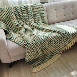 A textured throw blanket in shades of green, cream, and blue is draped over a light-colored sofa. The blanket features a bobble stitch design and a cream-colored fringe along the bottom edge. The blanket is made of a soft, cozy material.