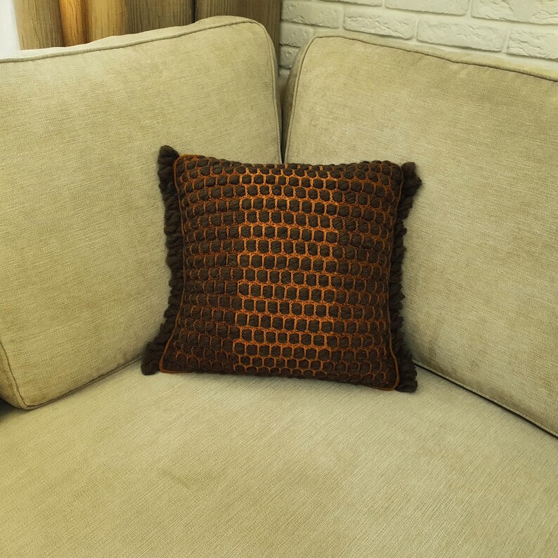Puede incluir: Un coj&iacute;n decorativo cuadrado con un patr&oacute;n texturizado en forma de panal en tonos marr&oacute;n y naranja. El coj&iacute;n tiene un fleco marr&oacute;n oscuro alrededor de los bordes y est&aacute; colocado sobre un sof&aacute; beige. Decoraci&oacute;n del hogar.