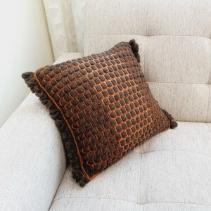 Puede incluir: Un coj&iacute;n decorativo cuadrado con un patr&oacute;n texturizado en forma de rejilla en tonos marr&oacute;n y naranja. El coj&iacute;n tiene un ribete de flecos en los bordes. El coj&iacute;n est&aacute; colocado sobre un sof&aacute; de color claro. Mide unos 40 cm x 40 cm.
