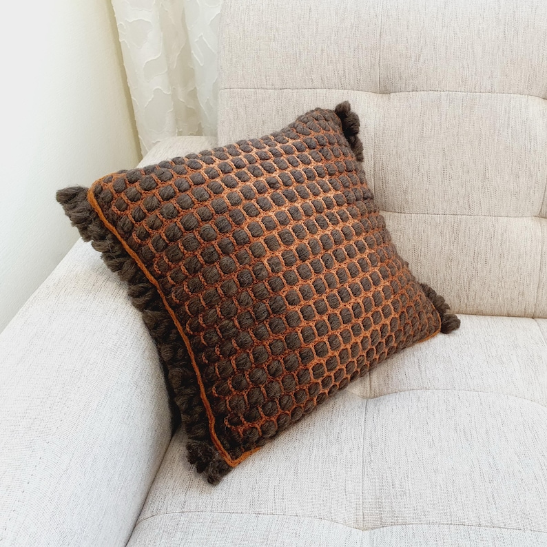 Puede incluir: Un coj&iacute;n decorativo cuadrado con un patr&oacute;n texturizado en forma de rejilla en tonos marr&oacute;n y naranja. El coj&iacute;n tiene un ribete de flecos en los bordes. El coj&iacute;n est&aacute; colocado sobre un sof&aacute; de color claro. Mide unos 40 cm x 40 cm.