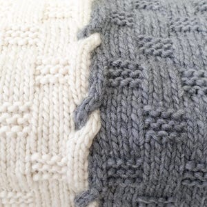 A close-up of a knitted throw blanket with a white and gray pattern. The blanket is made of soft yarn and has a textured surface.