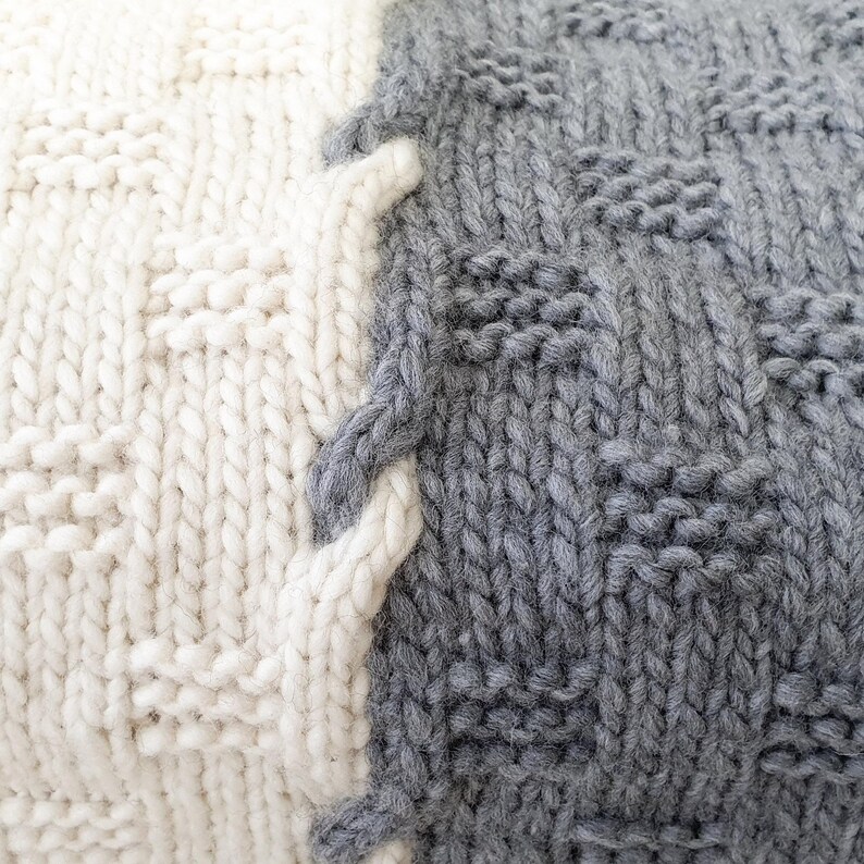 A close-up of a knitted throw blanket with a white and gray pattern. The blanket is made of soft yarn and has a textured surface.