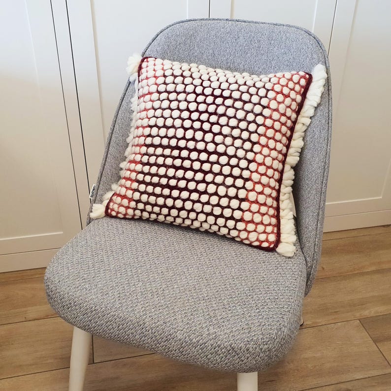 May include: A decorative square pillow with a textured white and burgundy pattern, fringed edges, and a white and burgundy color scheme. The pillow is placed on a gray chair with white legs.
