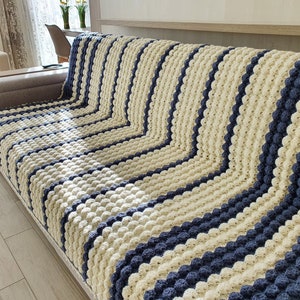 May include: A striped crocheted throw blanket in cream and navy blue. The blanket is made with a textured stitch that creates a raised, bumpy surface.