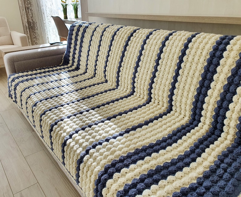 May include: A striped crocheted throw blanket in cream and navy blue. The blanket is made with a textured stitch that creates a raised, bumpy surface.