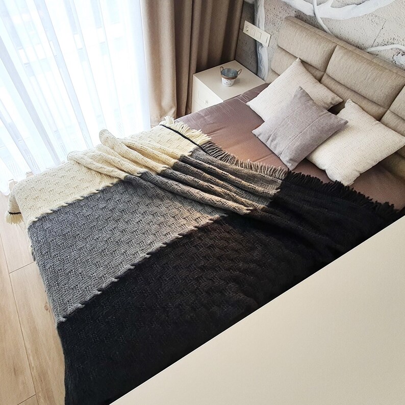 A bed with a white, gray, and black knitted throw blanket. The bed has a brown headboard and two pillows. The bed is in a bedroom with white curtains and a wooden floor.