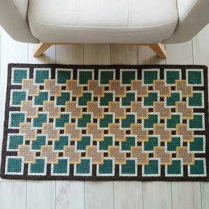 A brown and green geometric patterned rug with a square design. The rug is made of woven fabric and has a fringe border.