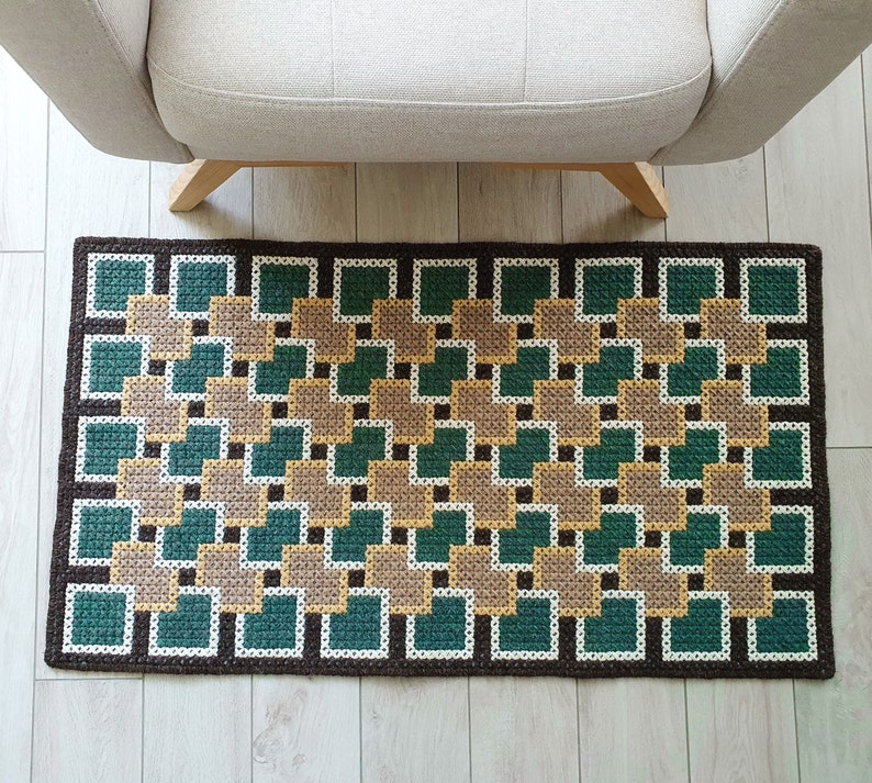 A brown and green geometric patterned rug with a square design. The rug is made of woven fabric and has a fringe border.