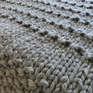 Close-up of a chunky knit blanket in a light gray color. The blanket features a textured pattern with raised, rounded stitches, creating a visually interesting surface. The thick yarn and tight weave suggest warmth and comfort.